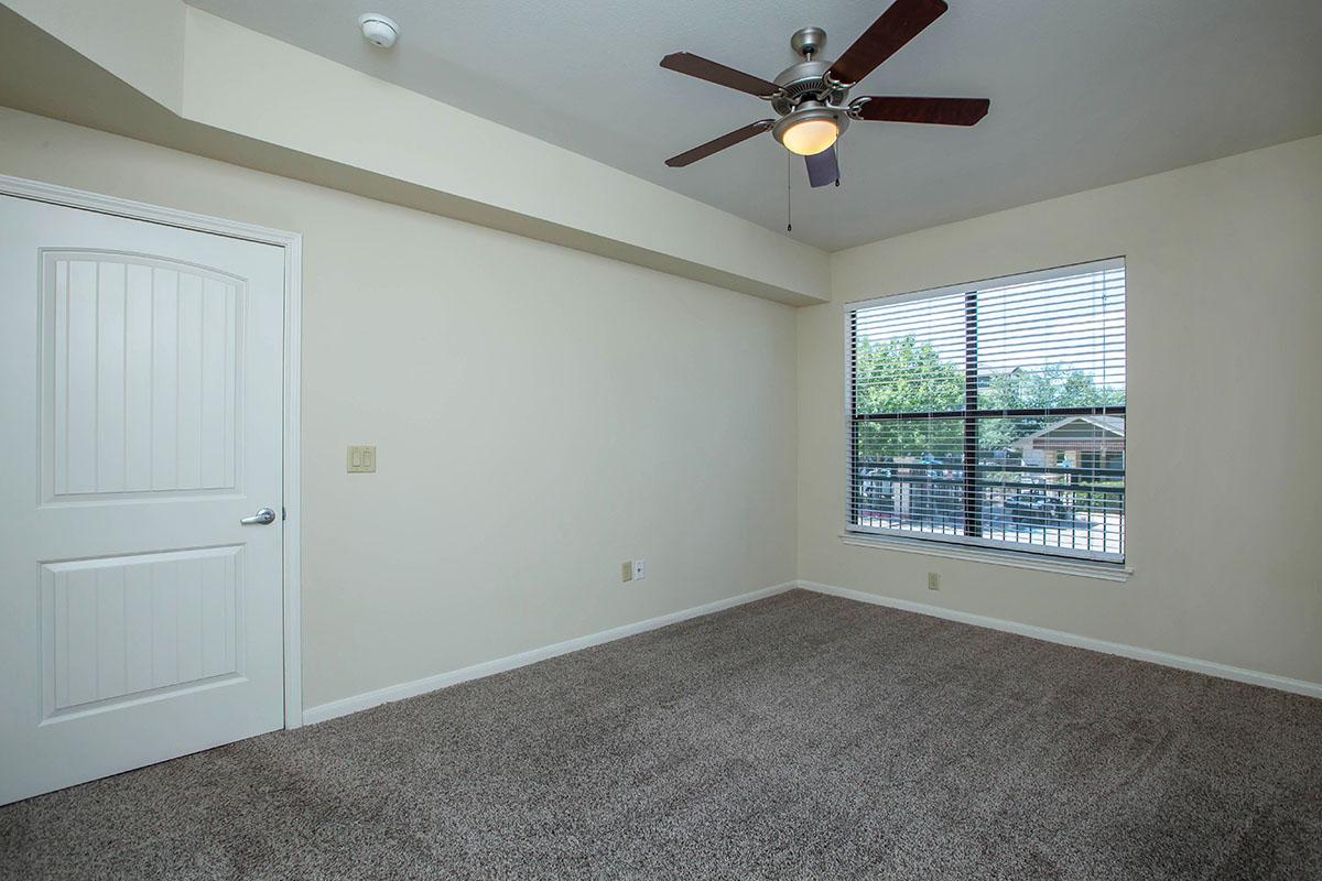 A well-lit, empty room with beige walls and carpet. It features a large window with blinds allowing natural light in, a ceiling fan, and a closed door. The space is clean and spacious, ready for furniture and decor.