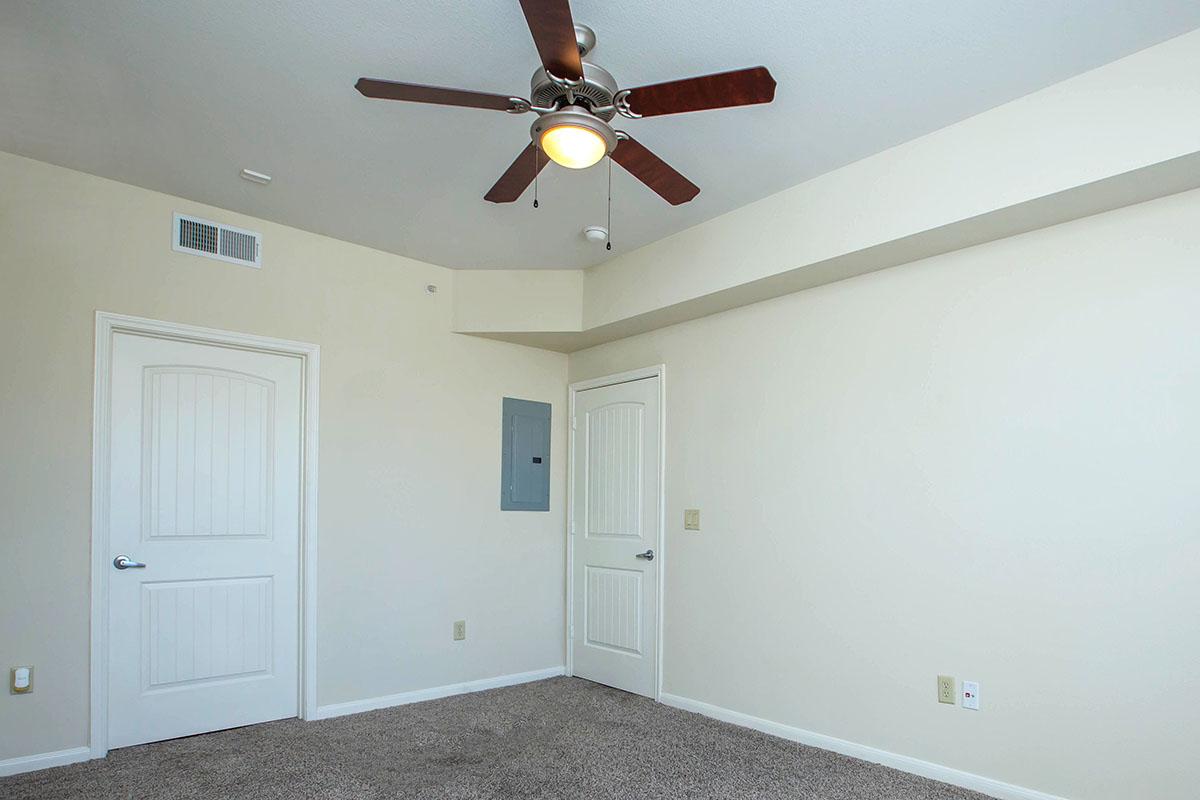 A brightly lit, unfurnished room with light beige walls and carpeted floor. It features a ceiling fan with wooden blades, a light fixture, and two doors—one leading to an adjoining room and the other possibly to an electrical panel. The room has a clean, minimalist look.