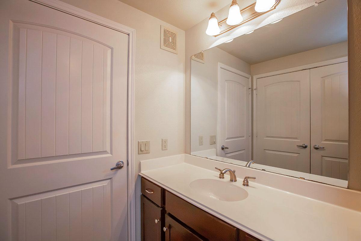 A well-lit bathroom featuring a white countertop with a sink, wooden cabinets underneath, a large mirror above, and a door leading to another room. The walls are painted a light color, and there are two closed doors visible in the background, enhancing the space's clean and modern look.