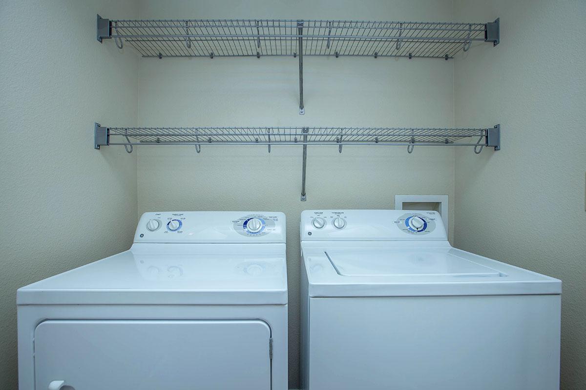 A laundry room featuring a white washer and dryer side by side. Above them, there are two metallic wire shelves mounted to the wall, providing storage space. The walls are a light neutral color, creating a clean and organized appearance.