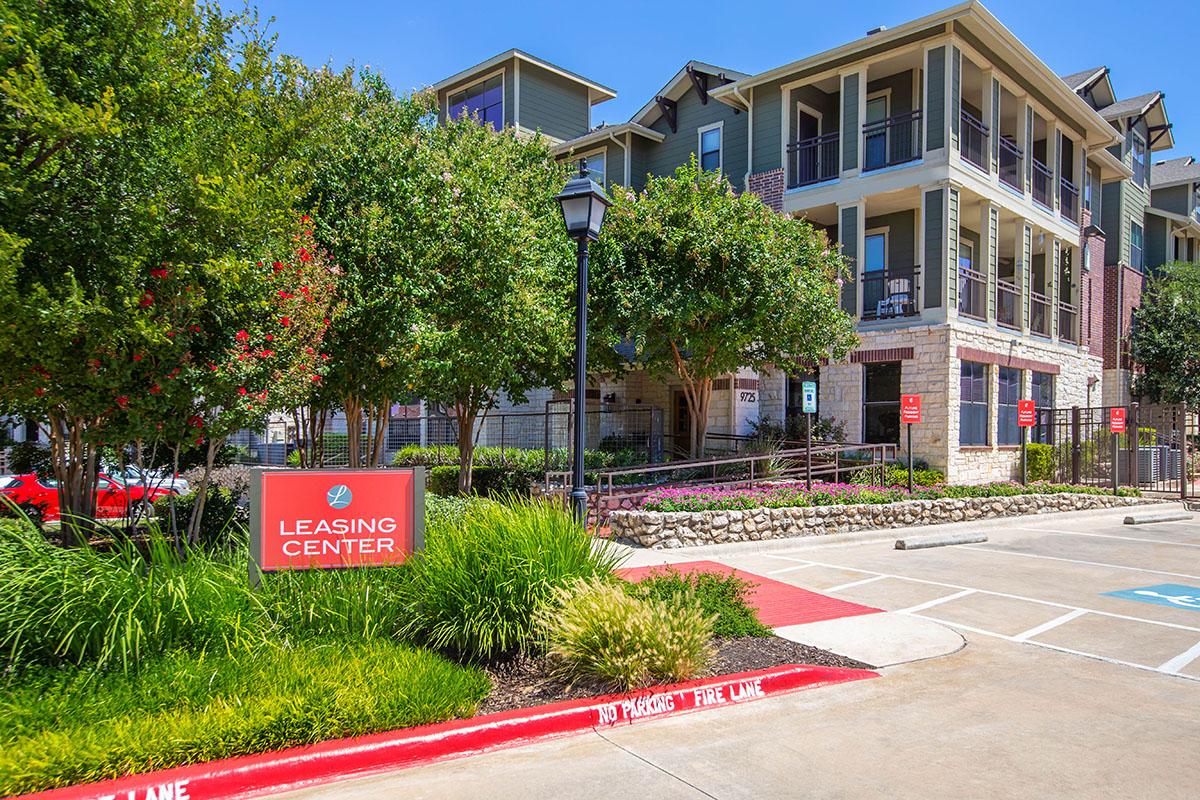 A leasing center for an apartment complex, featuring modern architecture with balconies and large windows. The area is landscaped with trees and shrubs, and a bold red sign indicates the leasing center's location. The driveway has clear markings and a 'No Parking Fire Lane' sign.