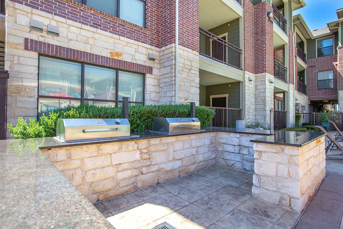 Outdoor grilling area featuring two stainless steel barbecue grills on a stone countertop. The scene includes a well-maintained apartment building with brick and stone facades, surrounded by greenery and outdoor seating.