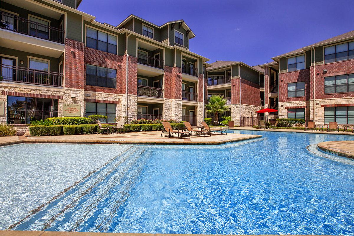 A clear swimming pool surrounded by lounge chairs, with brick apartment buildings in the background. The scene features lush greenery and bright blue skies, creating a relaxing outdoor environment.