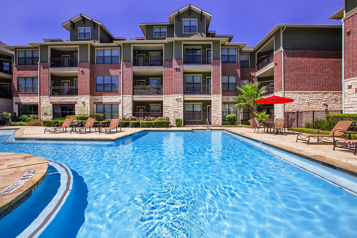A view of a spacious apartment complex featuring a blue swimming pool in the foreground. The pool is surrounded by lounge chairs, and there's a red umbrella offering shade. The building has a modern design with balconies, stone accents, and well-maintained landscaping under a clear blue sky.