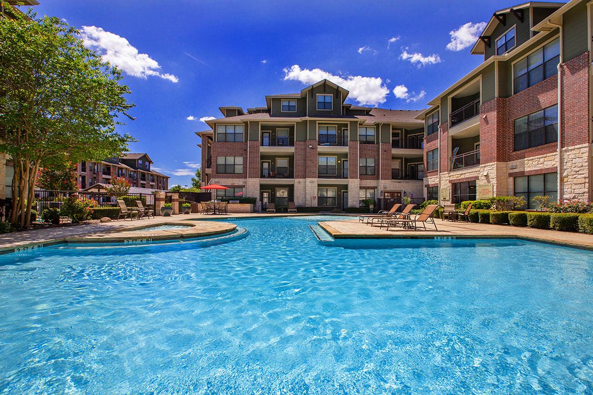A sunny apartment complex featuring a clear, blue swimming pool surrounded by lounge chairs and landscaped greenery. The buildings in the background have balconies and large windows, set against a bright blue sky with a few fluffy clouds.
