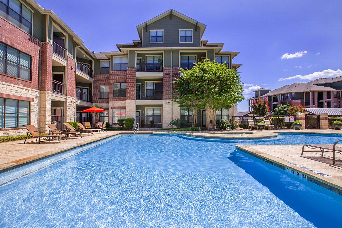 A clear swimming pool surrounded by lounge chairs and shaded umbrellas, set against an apartment building. The scene features vibrant greenery and a bright blue sky, creating a relaxing outdoor atmosphere ideal for leisure and social gatherings.