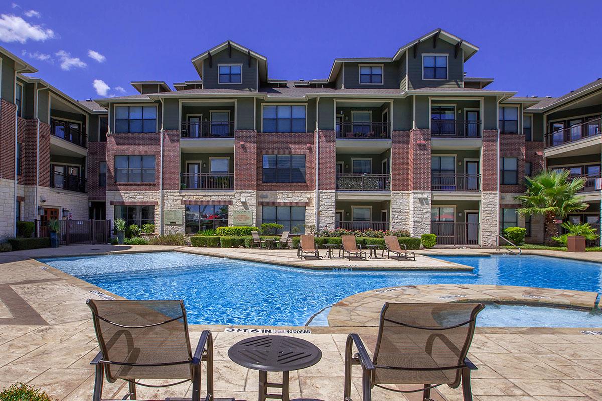 View of an outdoor swimming pool surrounded by lounge chairs, with a multi-story apartment building in the background. The building features a mix of brick and stucco architecture under a clear blue sky. Lush landscaping adds a vibrant touch to the area.