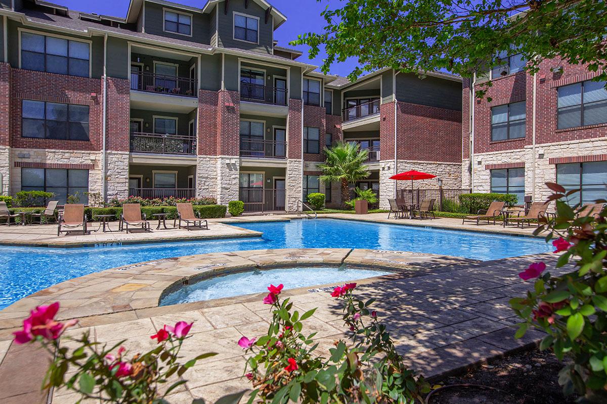 A sunny outdoor pool area featuring a swimming pool with a hot tub, surrounded by lounge chairs. In the background, there are multi-story apartment buildings with balconies. Brightly colored flowers are in the foreground, adding to the vibrant atmosphere of the space.