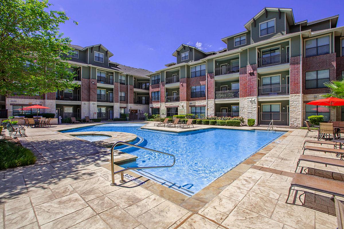 A sunny outdoor pool area surrounded by modern apartment buildings. The pool features a curved design with steps leading in, and red umbrellas dot the patio seating. Lush greenery enhances the inviting atmosphere, creating a relaxing environment for residents and guests.