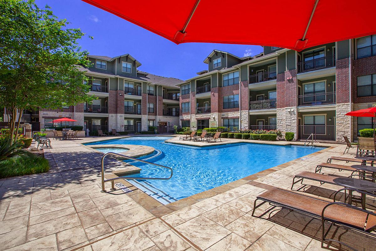A sunny courtyard featuring a large swimming pool surrounded by lounge chairs and shaded by red umbrellas. Modern apartment buildings are visible in the background, and neatly landscaped greenery adds to the inviting atmosphere. The clear blue sky enhances the vibrant setting.