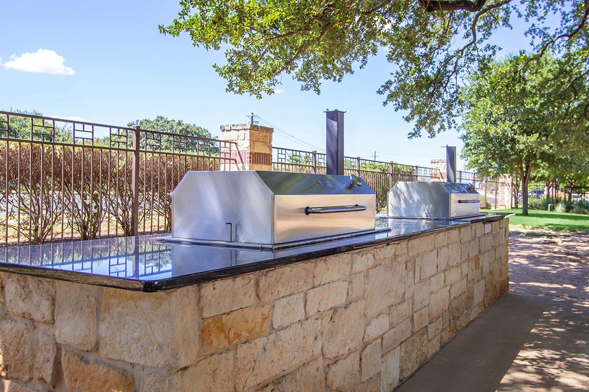 Two stainless steel grills with shiny surfaces are set on a stone countertop in an outdoor area. The grills are located near a fence, surrounded by greenery and trees under a clear blue sky with a few clouds. The setting appears inviting for barbecues and gatherings.