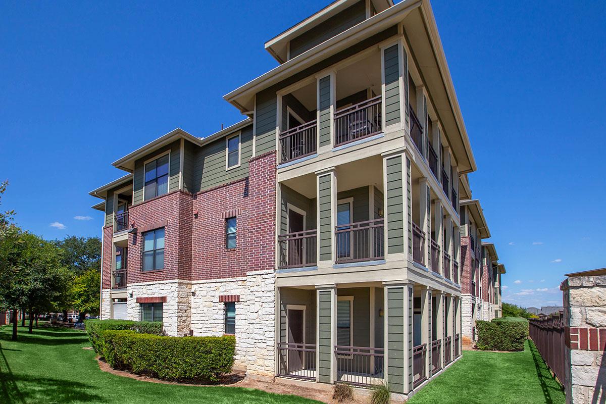 A multi-story apartment building featuring a combination of brick and siding exterior. The structure has several balconies on each floor, surrounded by well-maintained greenery and landscape. The sky is clear and blue, indicating a sunny day.