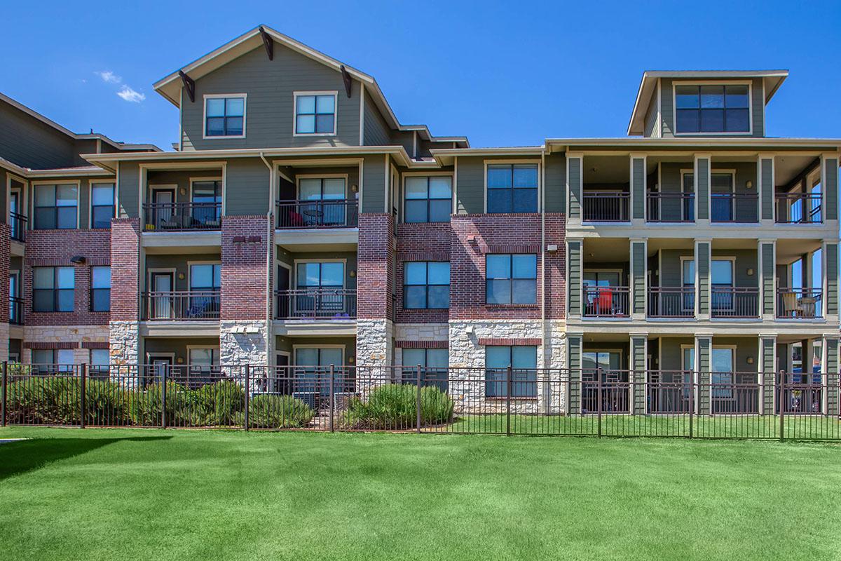 A modern apartment building featuring three stories with a mix of brick and stone exteriors. The structure has multiple balconies with sliding glass doors, set against a backdrop of a clear blue sky. The well-maintained lawn in the foreground adds greenery to the scene.