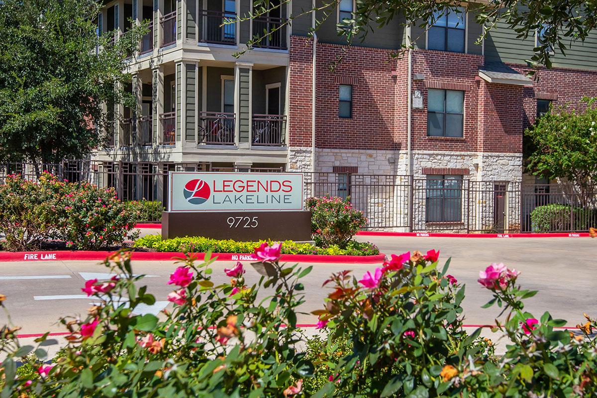 Sign for Legends Lakeline at 9725, surrounded by colorful blooming flowers and greenery, with a modern apartment building in the background.