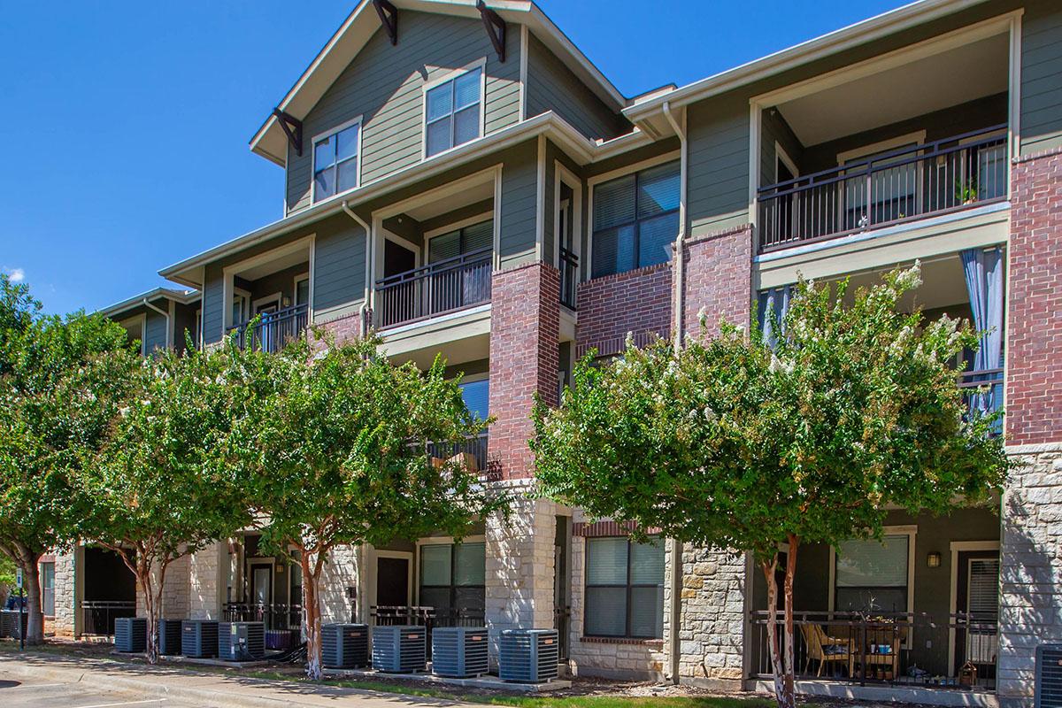A three-story apartment building featuring a mix of brick and siding exteriors. It has balconies with railings on each floor and is surrounded by neatly trimmed trees. Air conditioning units are visible on the ground level. The sky is clear and blue, enhancing the appearance of the property.