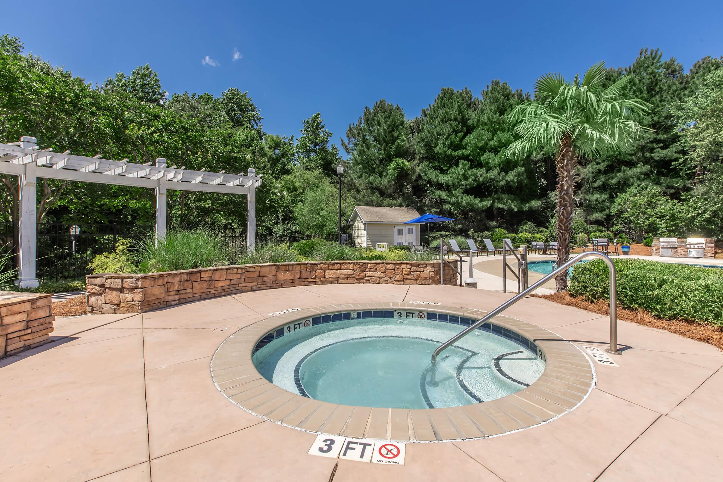 A sunny outdoor area featuring a circular hot tub surrounded by a stone patio, tropical plants, and a pergola. In the background, there are lounge chairs and a swimming pool. Signs indicate the hot tub's depth is 3 feet. Lush greenery surrounds the space, creating a relaxing atmosphere.