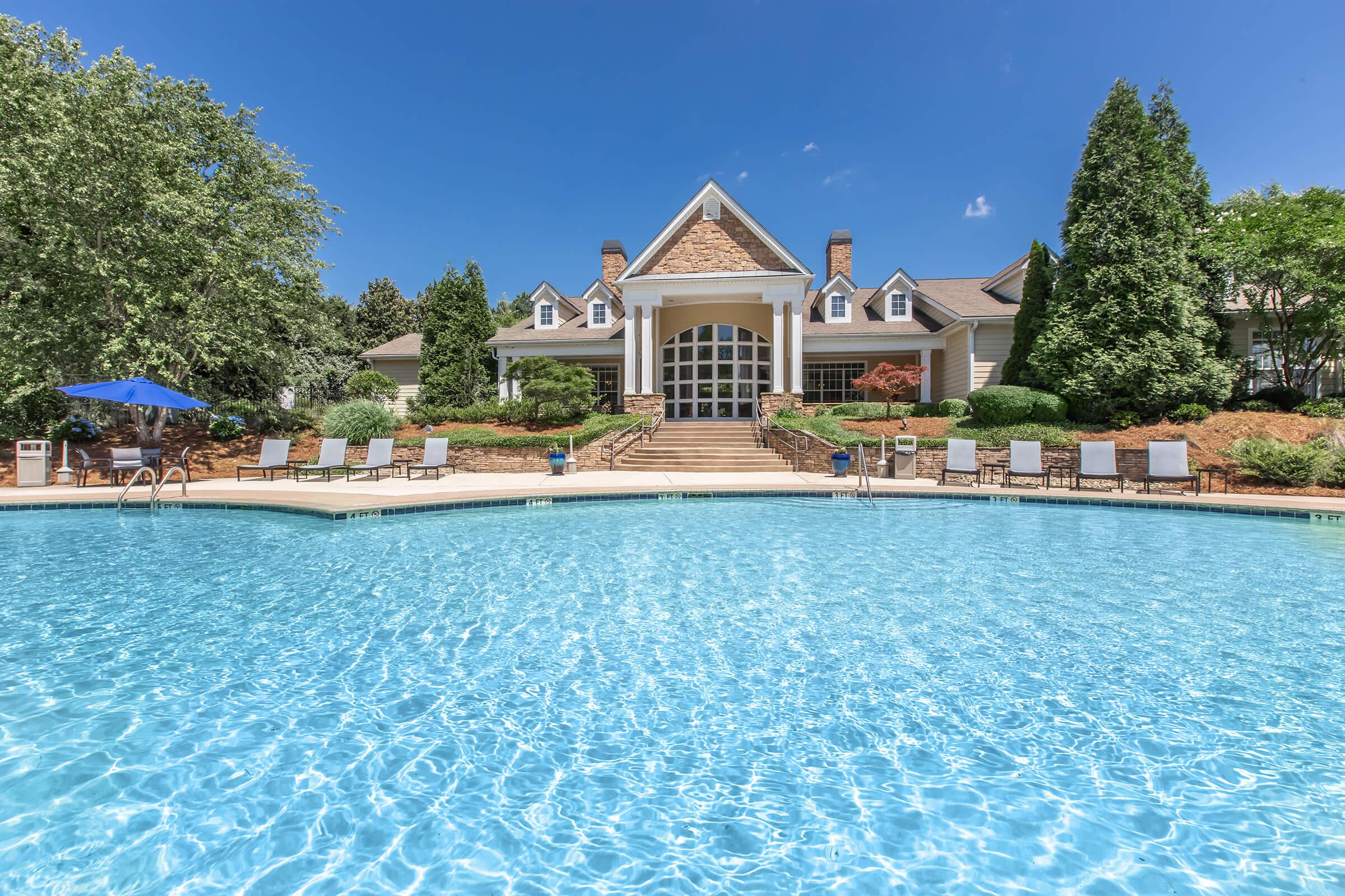 A sparkling blue swimming pool in the foreground, surrounded by lounge chairs. In the background, a large, elegant building with a stone facade and large windows is framed by lush greenery and trees under a clear blue sky. The scene conveys a calm and inviting atmosphere.