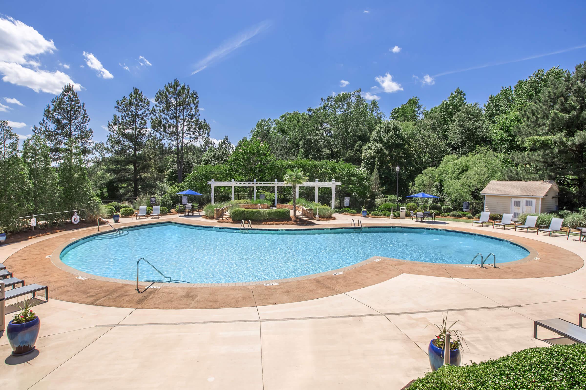 A serene outdoor swimming pool surrounded by lush green trees and shrubs. The pool, with a curved shape and clear blue water, features lounge chairs and umbrellas nearby. A covered pavilion stands in the background, adding to the tranquil atmosphere of the setting. Bright blue sky with a few clouds overhead.