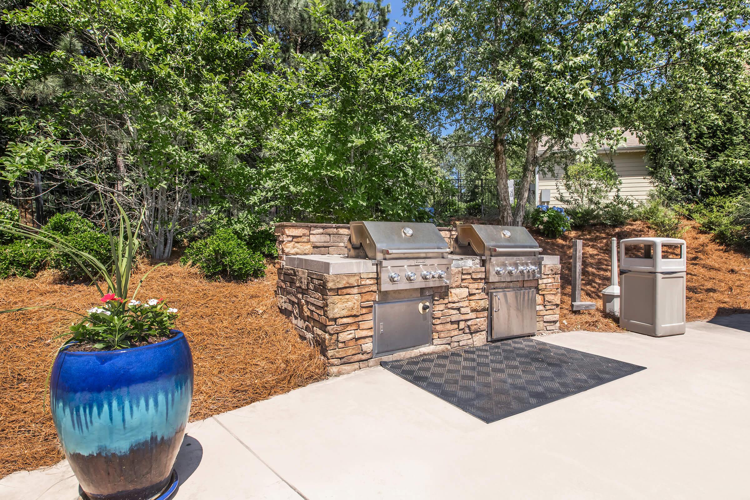 An outdoor grilling area featuring two stainless steel grills set on a stone base, surrounded by lush greenery and a landscaped area with mulch. A blue decorative planter with flowers is visible alongside the grilling station, which includes a black mat and a trash bin nearby.