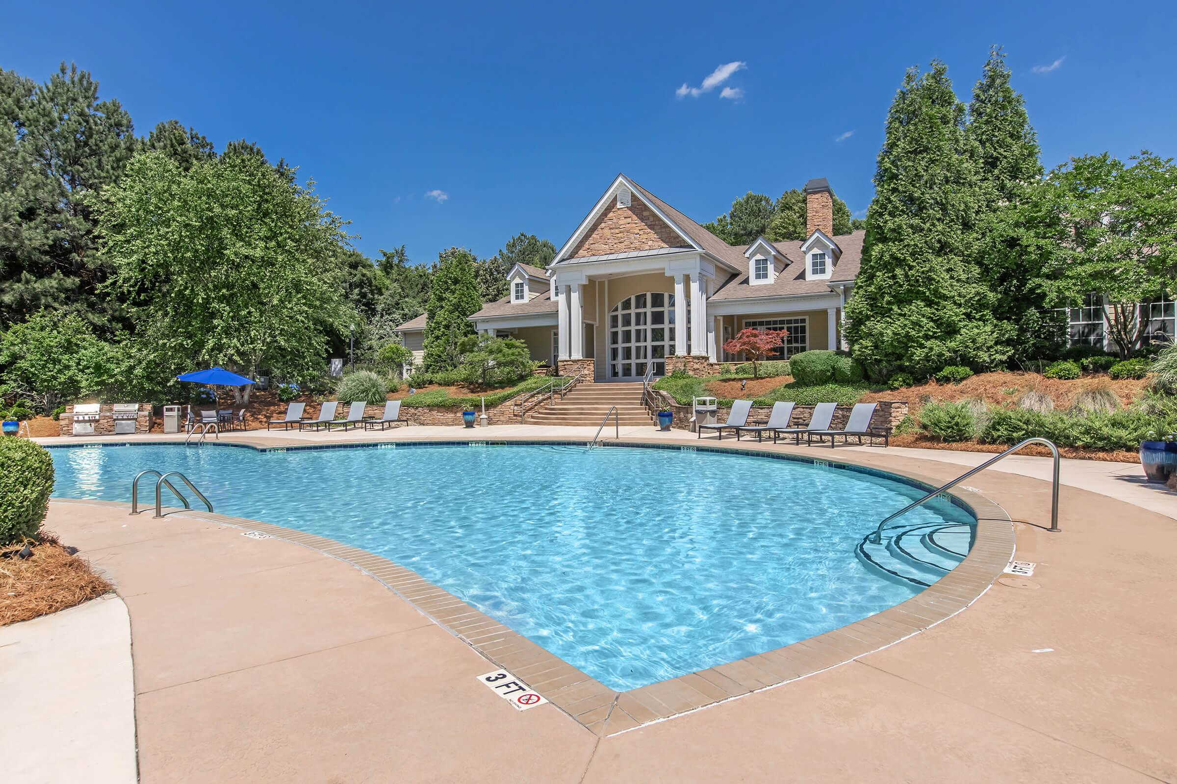 A large outdoor swimming pool with clear blue water, surrounded by lounge chairs and lush greenery. In the background, there's a well-maintained building featuring a classic architectural style. The sky is bright blue with a few clouds, creating a serene and inviting atmosphere.