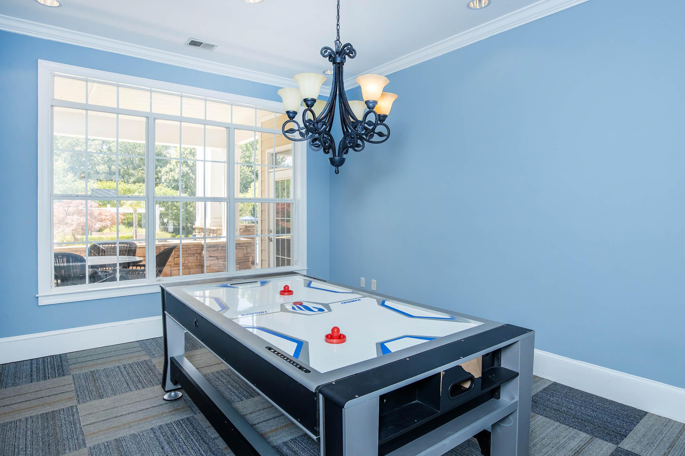 A light blue room featuring an air hockey table with red paddles and a chandelier hanging from the ceiling. Large windows let in natural light, revealing an outdoor area visible through the glass. The floor is covered with carpet tiles in a muted pattern.