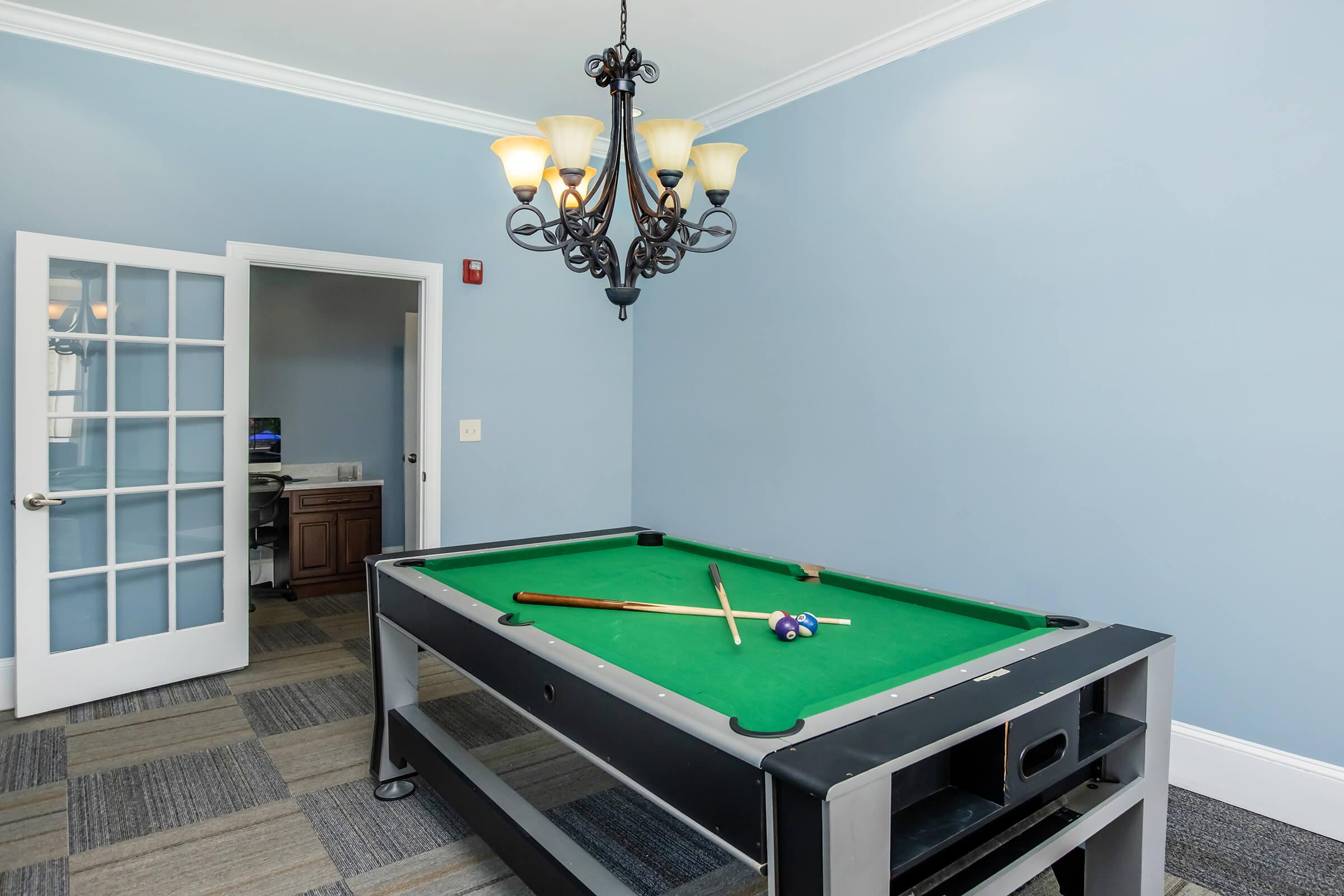 A billiards table with a green felt surface and cue sticks rests in a well-lit room featuring blue walls and a chandelier. A set of pool balls is scattered on the table, and a doorway leads to another room in the background. The flooring is a mix of patterned tiles.