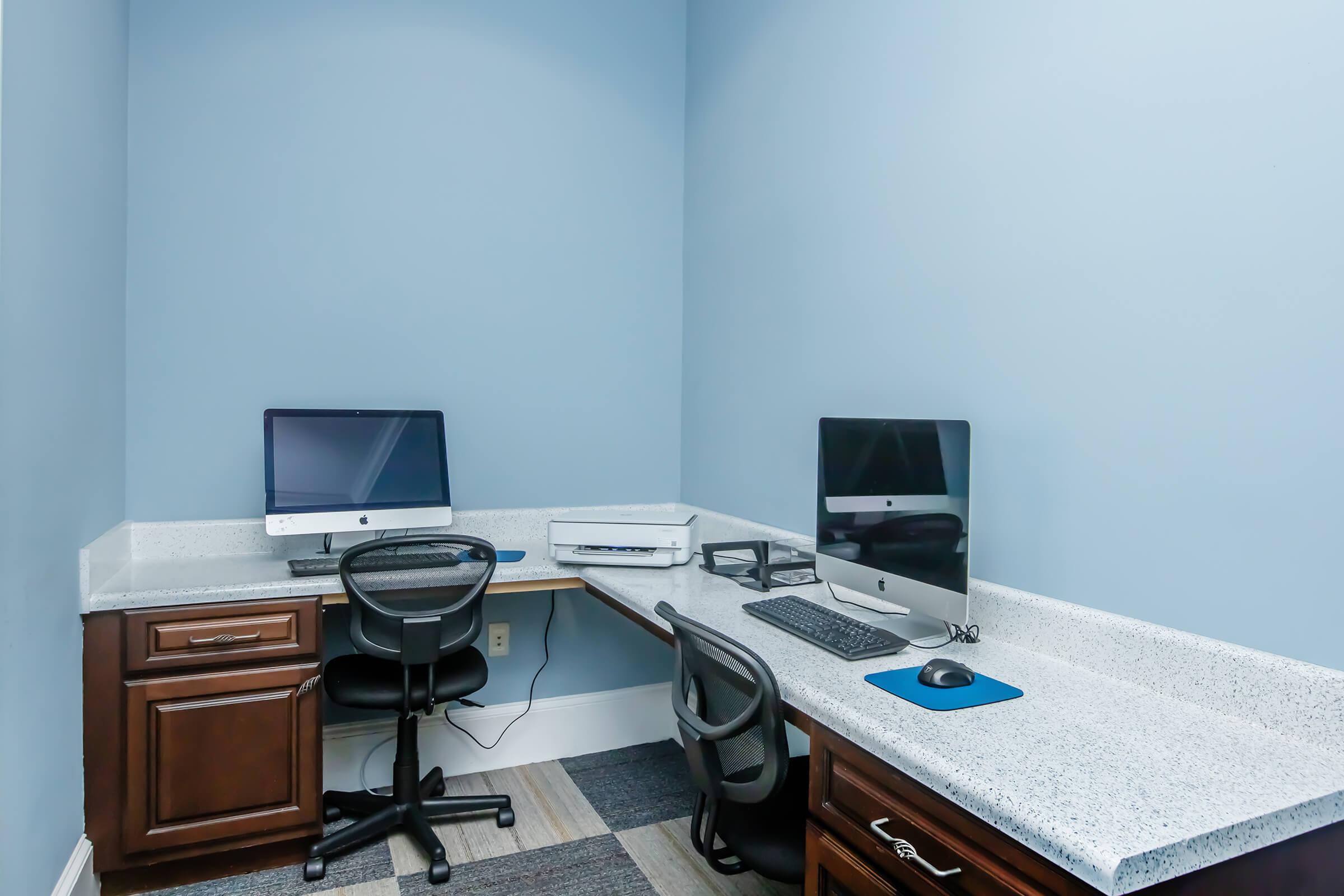 A modern office space featuring two computer workstations with iMacs, ergonomic chairs, and a printer. The walls are painted a light blue, and the desks have a sleek, white countertop. The room has a clean, organized appearance, suitable for productivity.