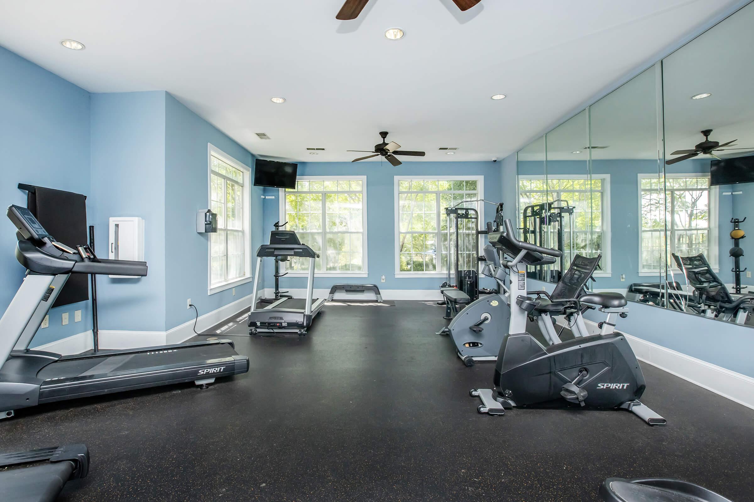 A modern gym interior featuring various fitness equipment, including treadmills, stationary bikes, and weight machines. The space has large windows allowing natural light, creating a bright and inviting atmosphere, and mirrors on one wall that enhance the feeling of openness.