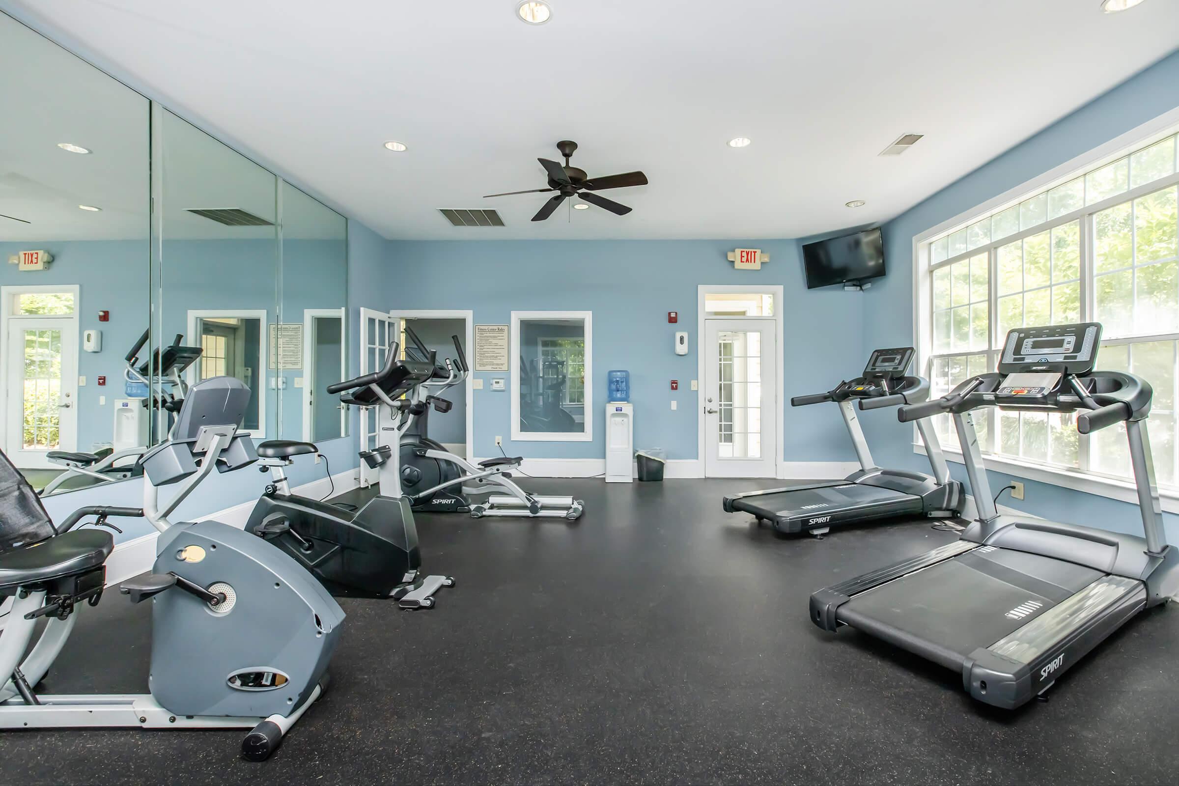 A well-lit gym area with exercise equipment including two treadmills, a stationary bike, and a multi-station weight machine. Large windows provide natural light, while mirrors on one wall enhance the space. A ceiling fan adds comfort, and a water cooler is visible in the background.