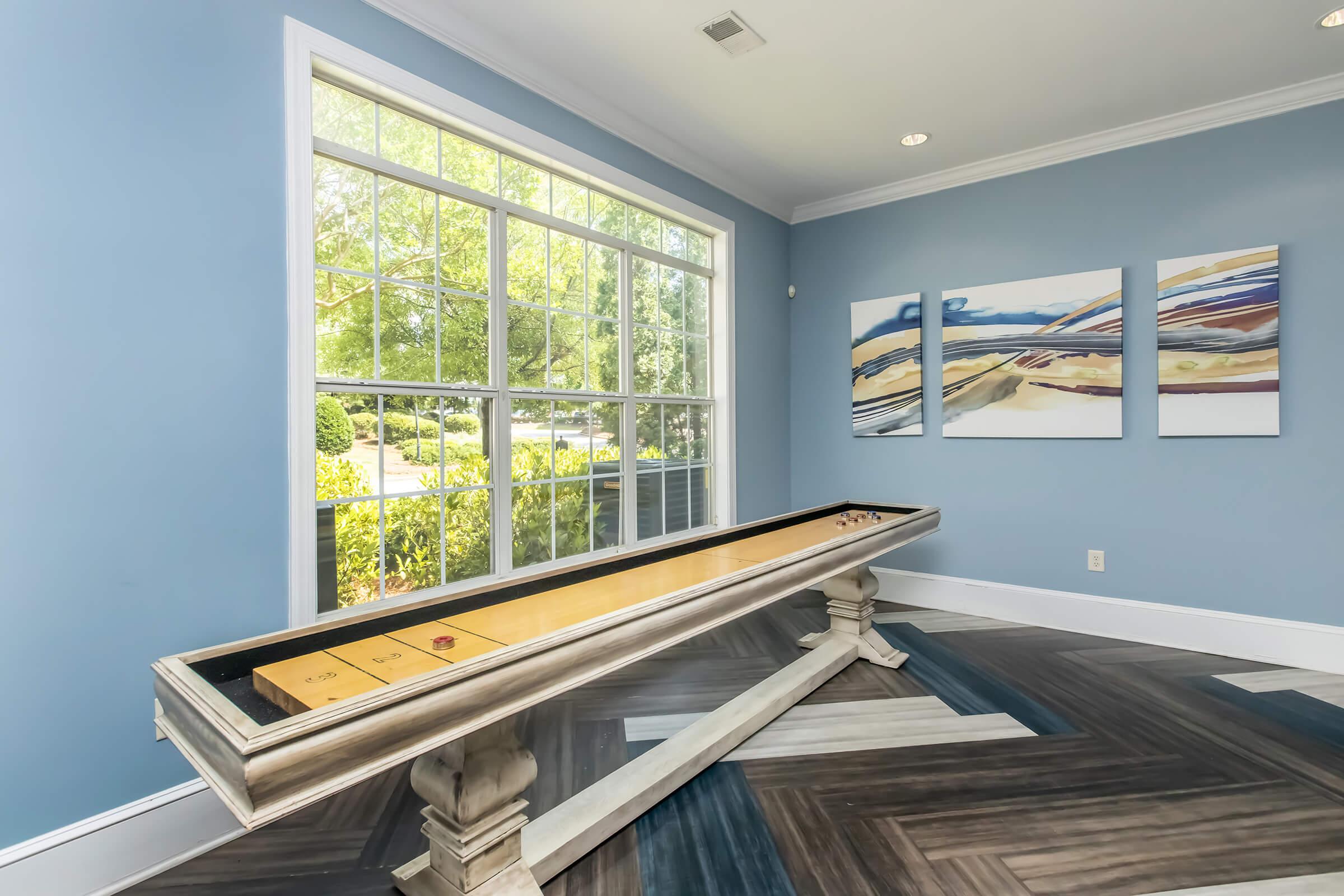 A stylish game room featuring a long shuffleboard table, positioned against a large window that lets in natural light. The walls are painted light blue, and there are three abstract paintings in soft colors hanging nearby. The floor features a modern herringbone pattern.