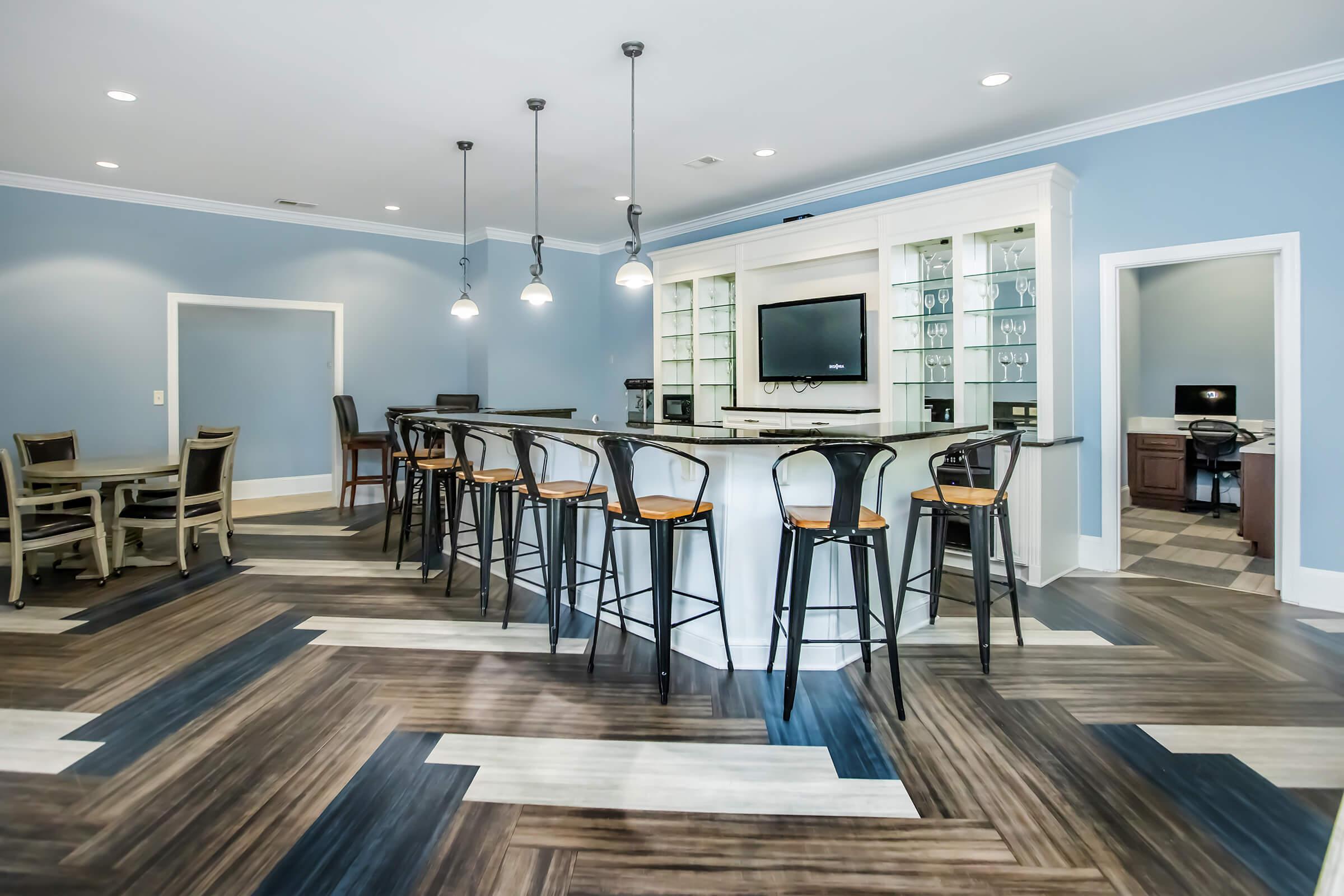 A modern lounge area featuring a large bar with stools, a television mounted on the wall, and a mix of wood flooring. Light blue walls complement the sleek design, which includes a dining table and chairs in the background. Stylish pendant lights hang above the bar, creating an inviting atmosphere.