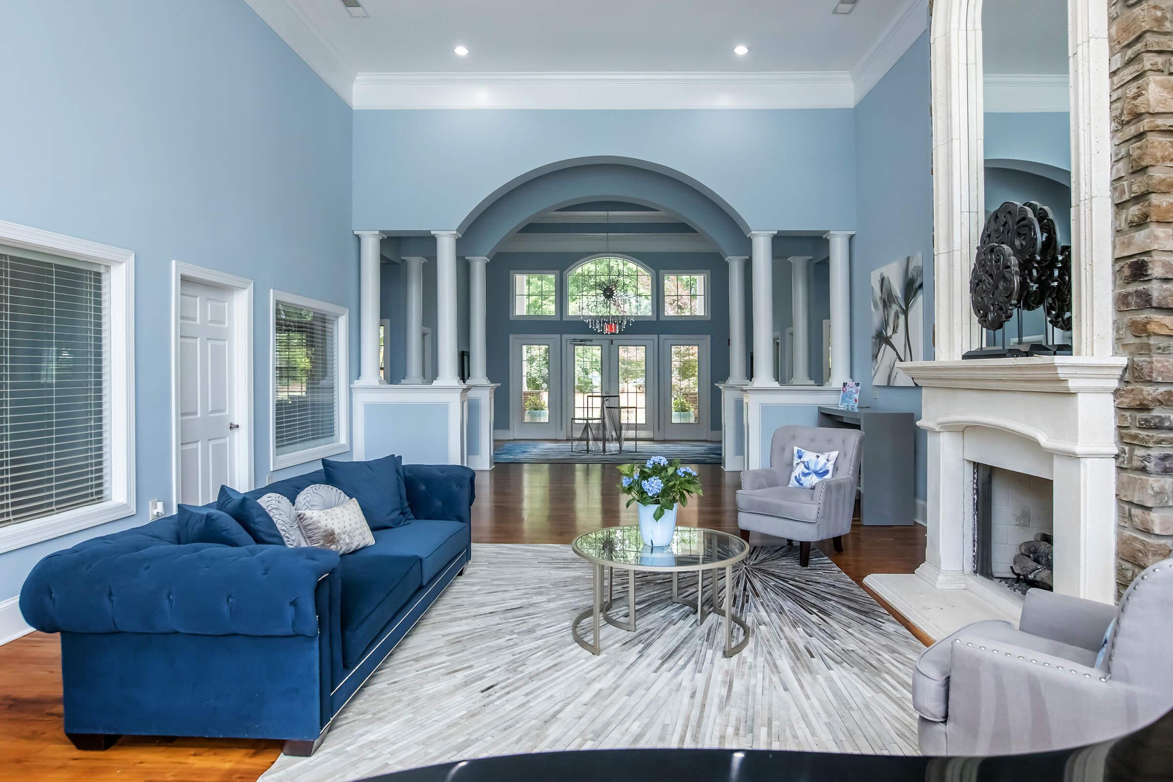 Spacious living room with blue walls and large windows, featuring a plush blue sofa, a round glass coffee table with a floral arrangement, and a cozy chair. A stone fireplace is visible, along with hardwood floors, creating an inviting and elegant atmosphere.