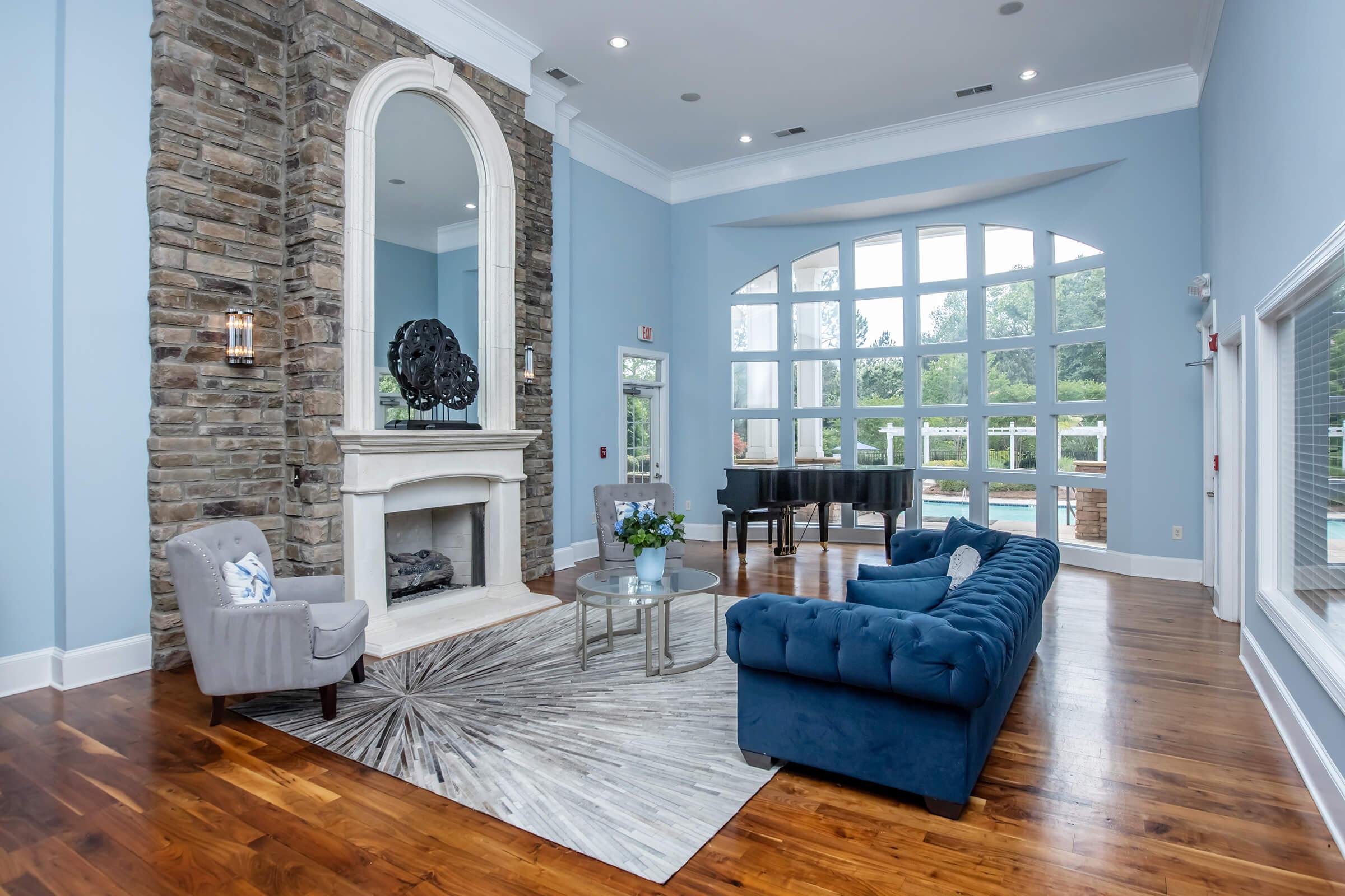 A spacious living room featuring a stylish blue velvet sofa, a round glass coffee table, and decorative elements. The room has a large stone fireplace with a mirror above it, and floor-to-ceiling windows allowing natural light. A piano is in one corner, with a view of the outdoor pool visible through the windows.