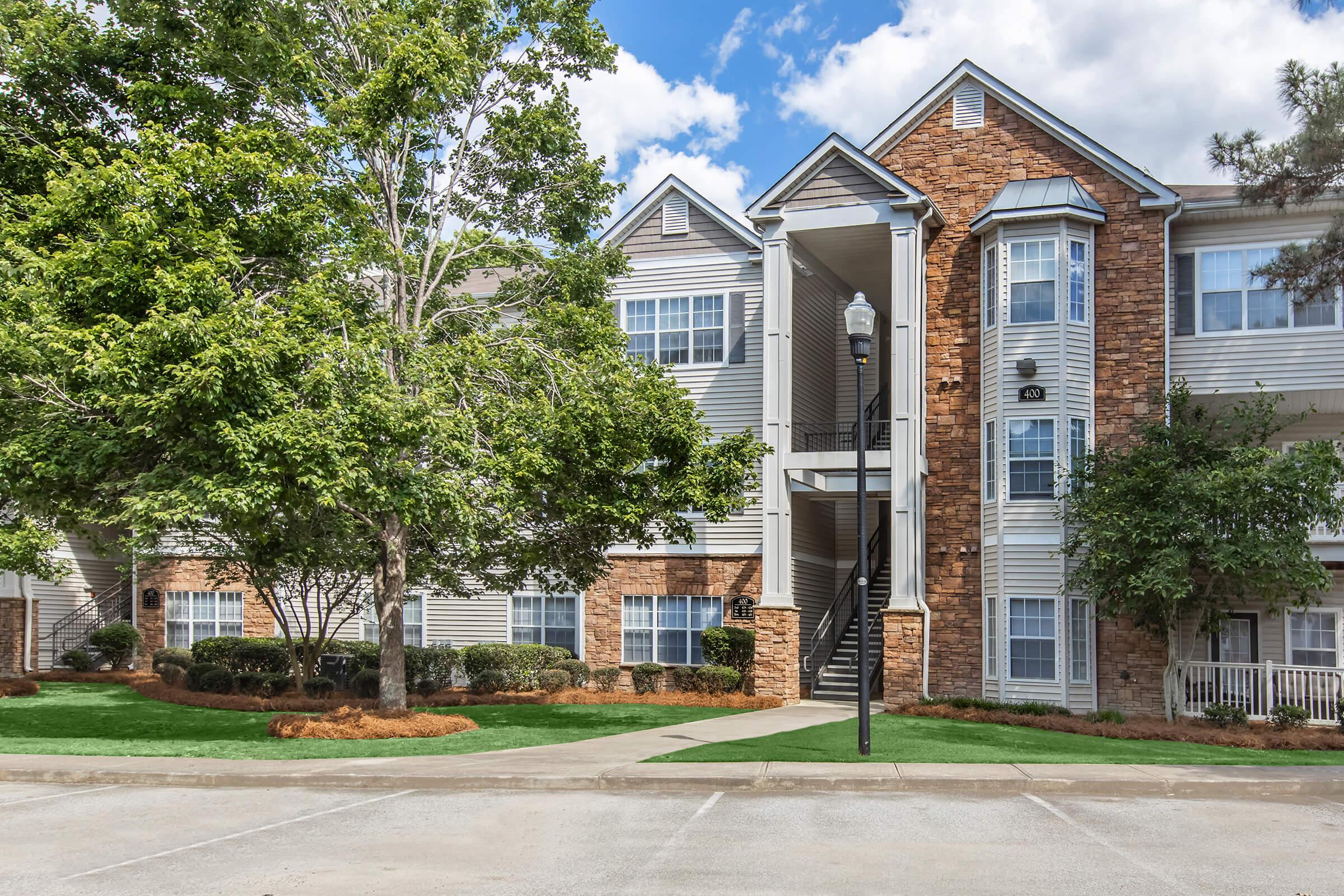A multi-story apartment building featuring a mix of brick and siding exteriors. Lush greenery surrounds the building, with a well-maintained lawn. Stairs lead up to the entrance, and the sky above is partly cloudy, contributing to a bright and inviting atmosphere.