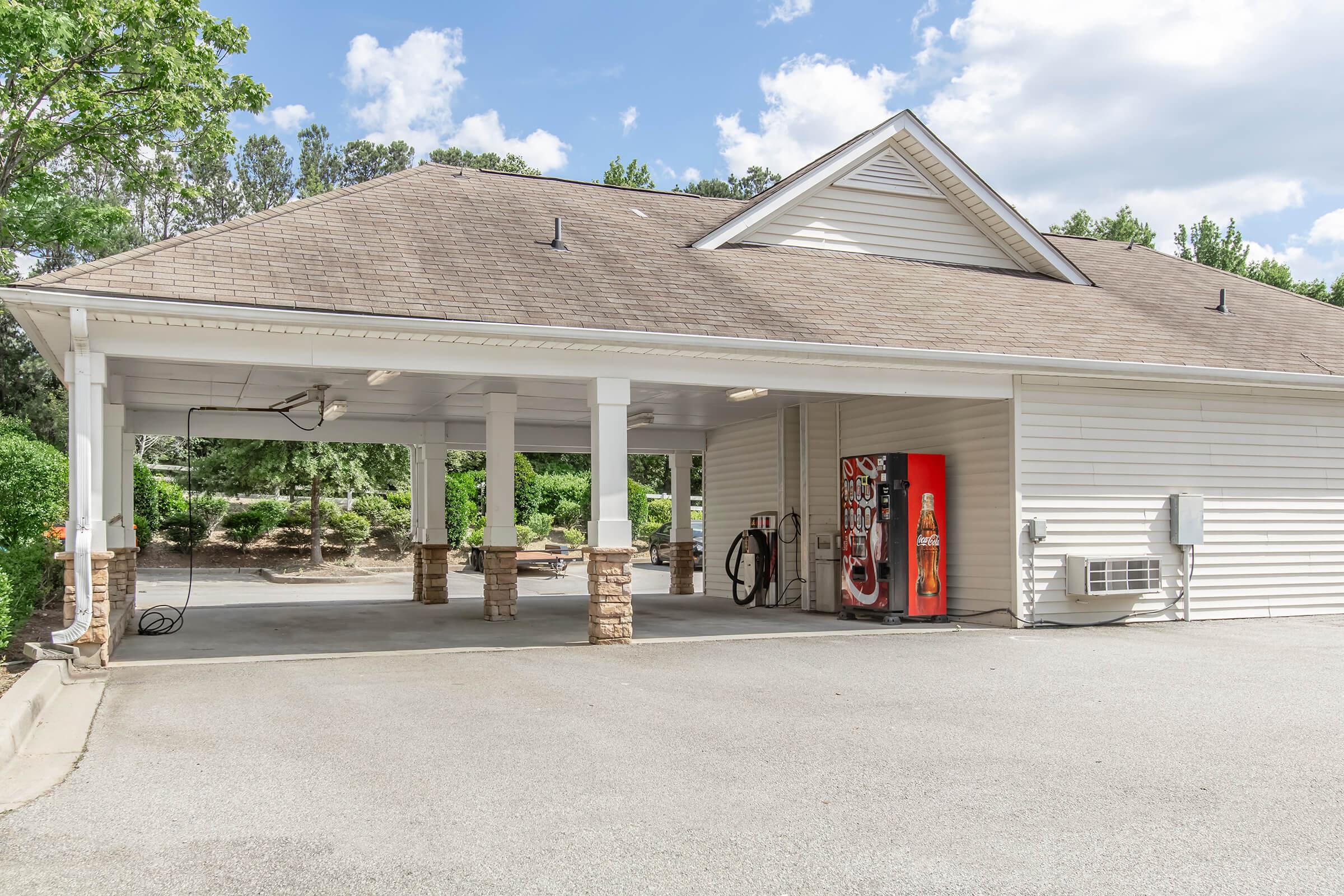 A covered parking area with stone pillars, featuring a vending machine on one side, surrounded by greenery and trees. The structure has a sloped roof with a light color and is set against a bright sky with clouds.