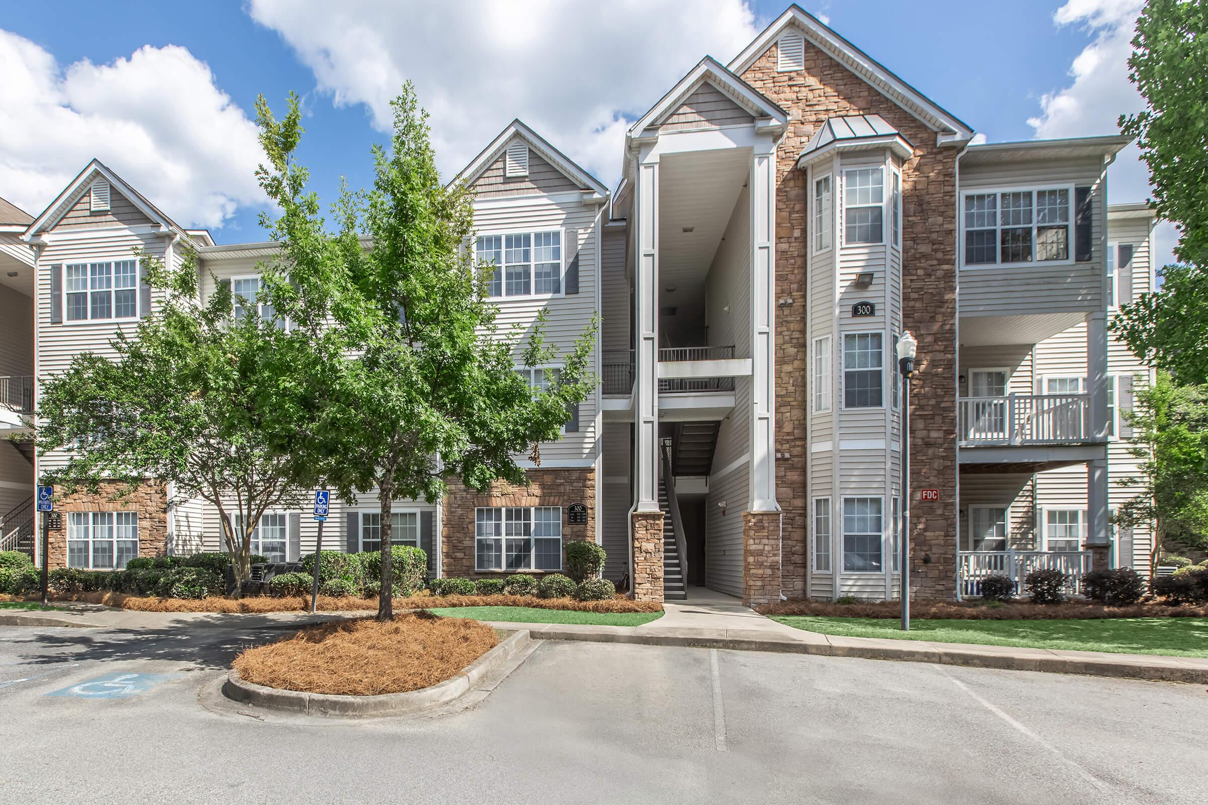 Exterior view of a modern multi-story apartment building featuring a combination of brick and siding. Lush green landscaping surrounds the building, with parking in the foreground. The sky is partly cloudy, creating a bright and inviting atmosphere.