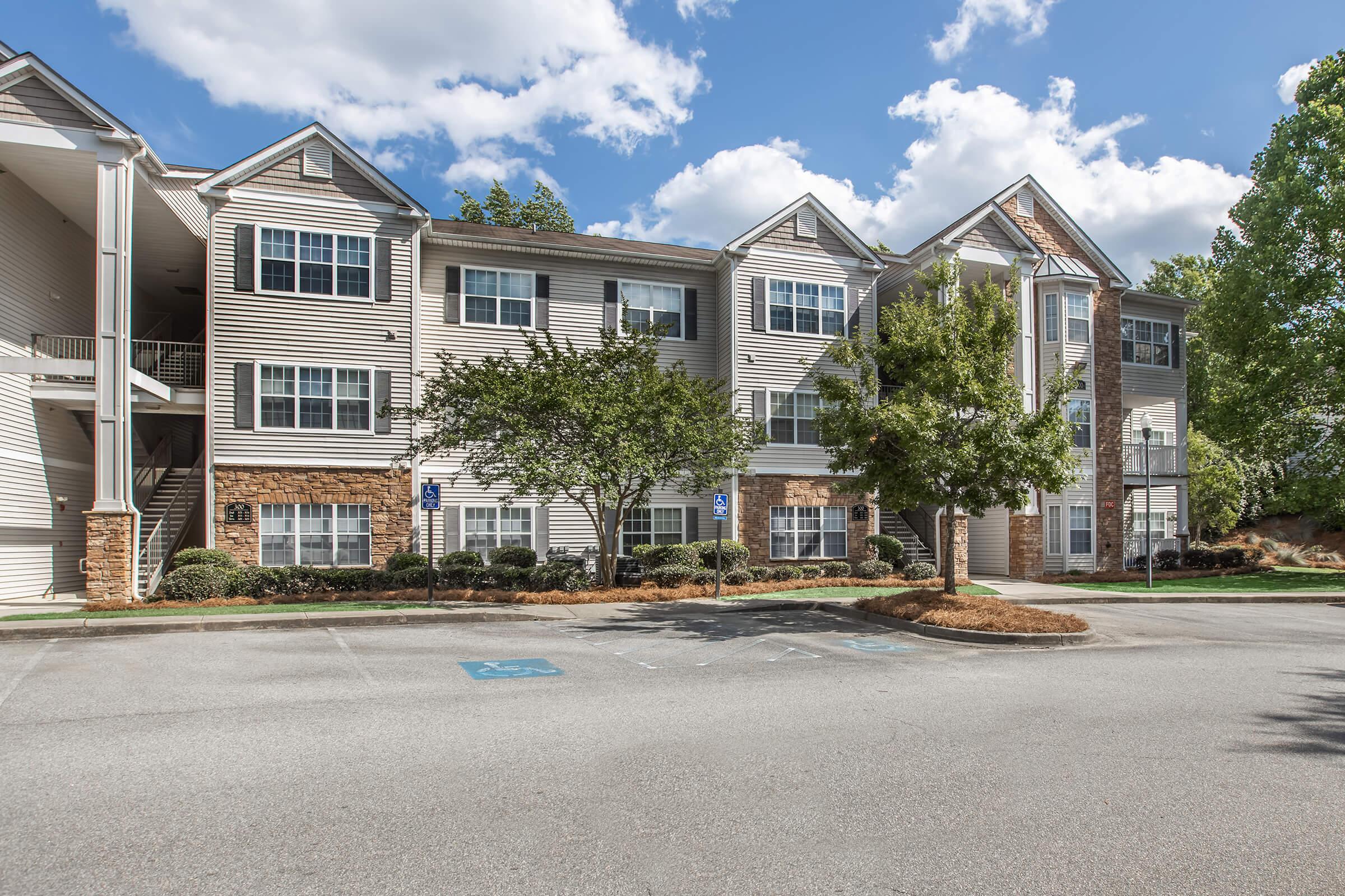 Exterior view of a residential apartment building with multiple stories, featuring a mix of siding and stone elements. Surrounding greenery includes trees and shrubs. The parking area shows designated spaces, and there are clear blue skies with white clouds overhead.