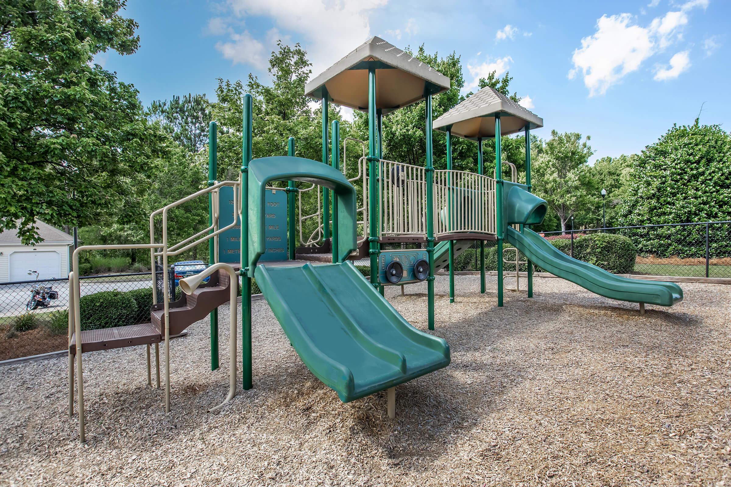 A playground featuring two green slides, a climbing structure with multiple levels, and safety fencing. Surrounding the playground are trees and a grassy area, with a blue sky overhead. Mulch covers the ground beneath the equipment for safety.