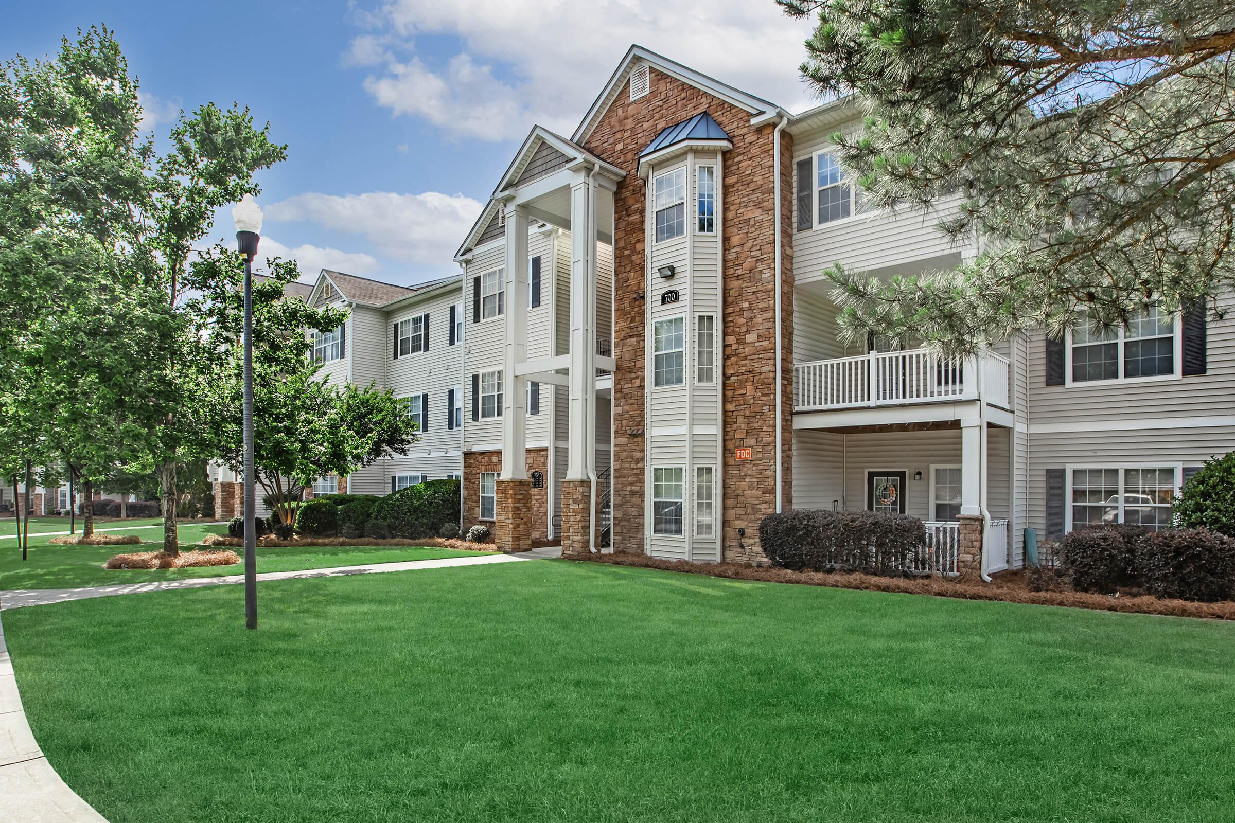 A spacious two-story residential building with a mix of white siding and stone accents, surrounded by well-maintained green lawns, shrubs, and trees. The building features multiple balconies and a central entrance with decorative pillars leading to the main door.