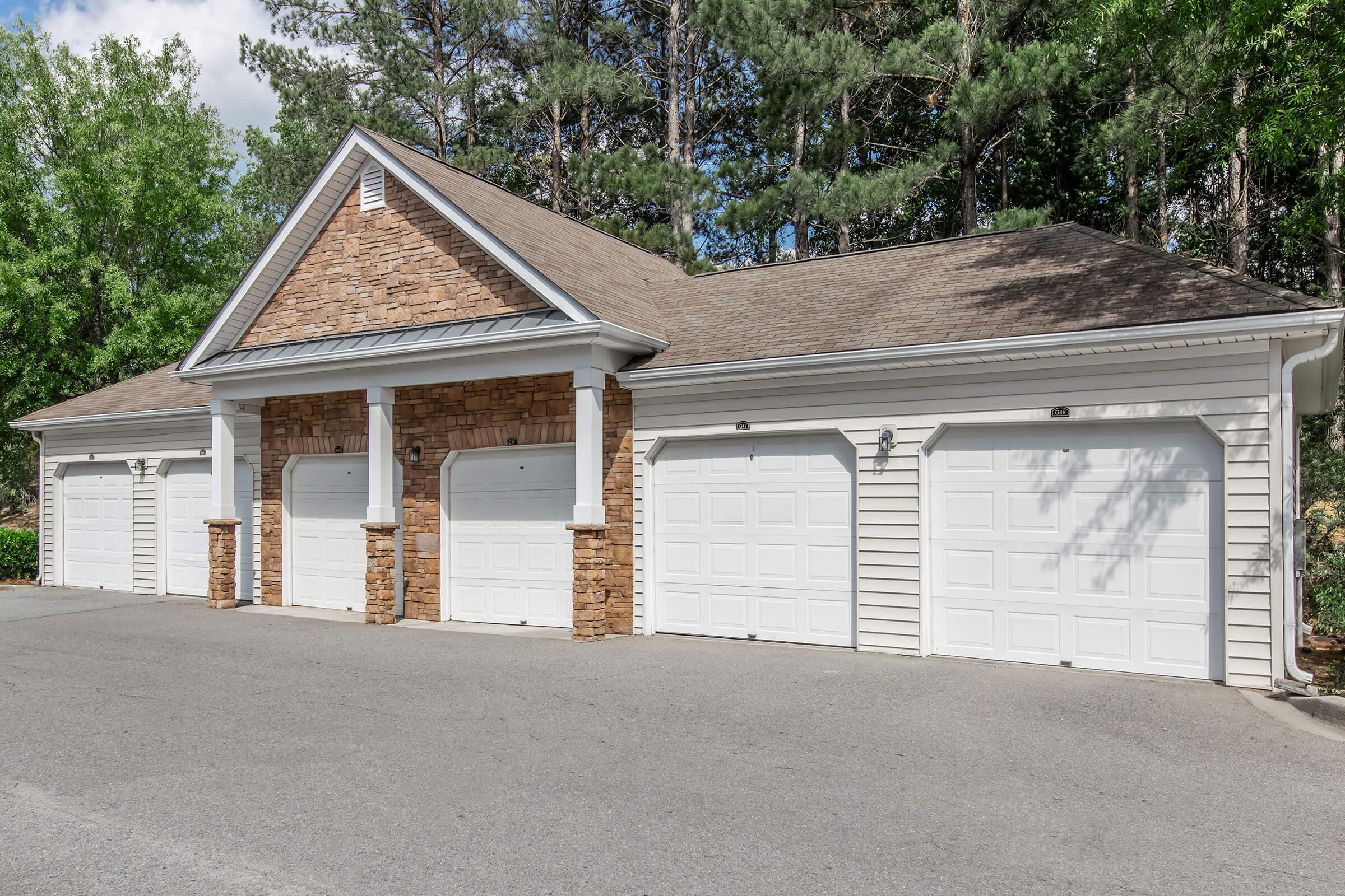 A modern garage structure with three white garage doors, flanked by tan siding and stone accents. The building features a gabled roof and decorative columns at the entrance, set against a backdrop of green trees and a clear blue sky. The driveway is paved and well-maintained.