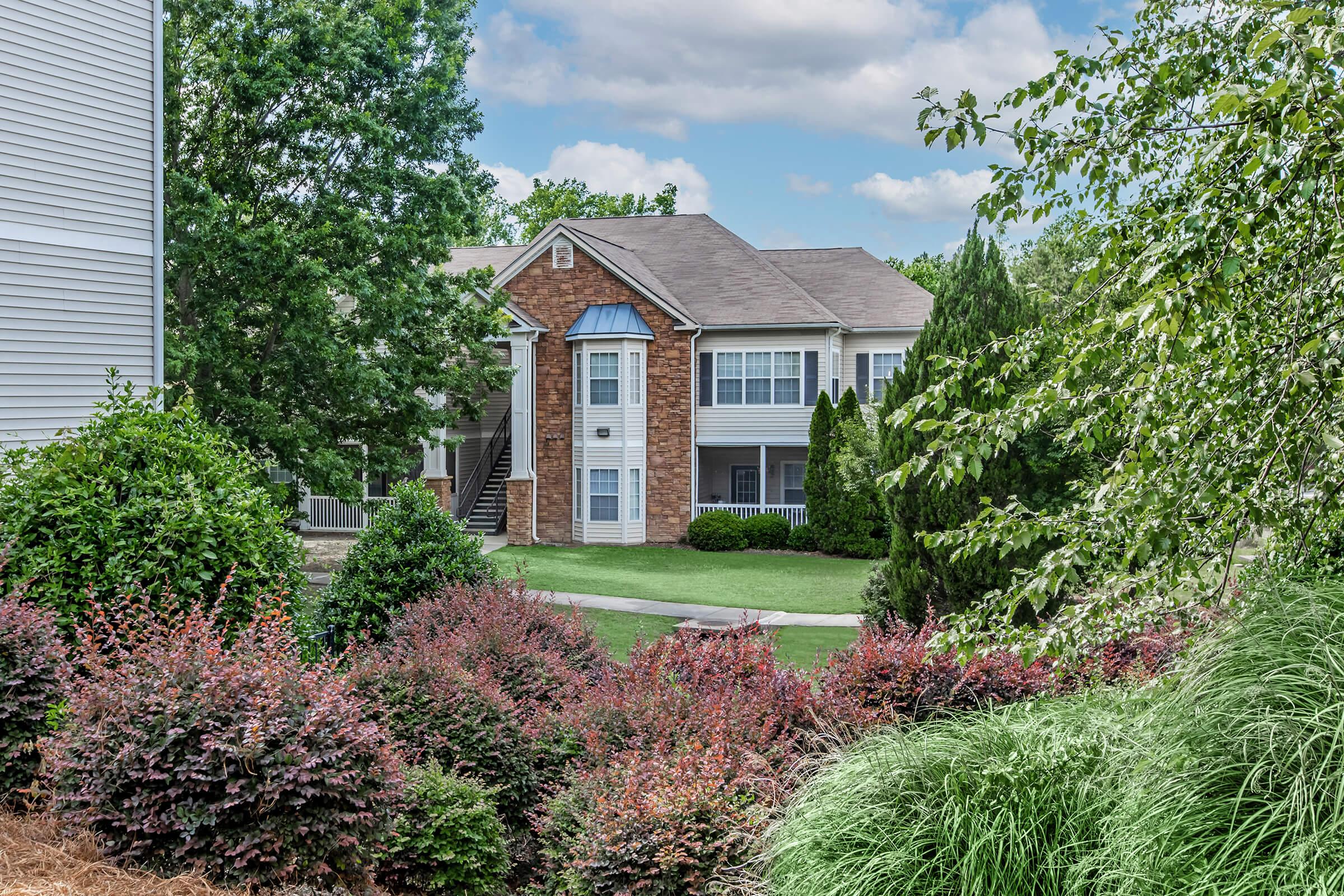 A residential building with a mix of brick and siding, surrounded by well-maintained landscaping featuring green bushes and vibrant flowering plants. The scene includes a grassy area, trees, and a clear sky, suggesting a serene suburban atmosphere.