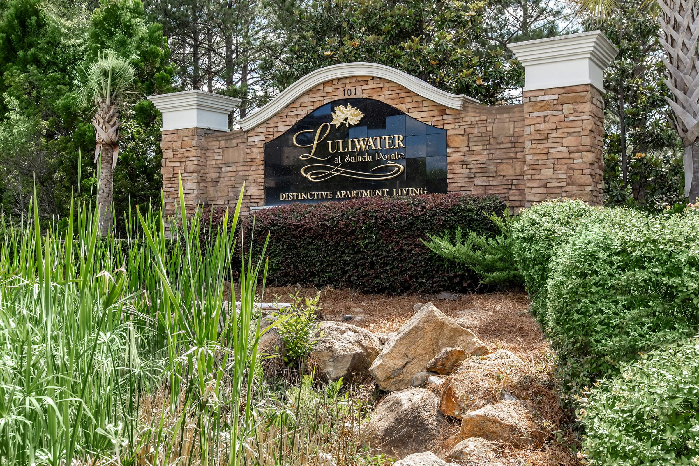 Sign for Lullwater at Ashford Pointe, an apartment community. The sign features the name "Lullwater" prominently and includes the phrase "Distinctive Apartment Living." Surrounded by lush greenery, including tall grass and trimmed bushes, the image reflects a well-maintained landscape.