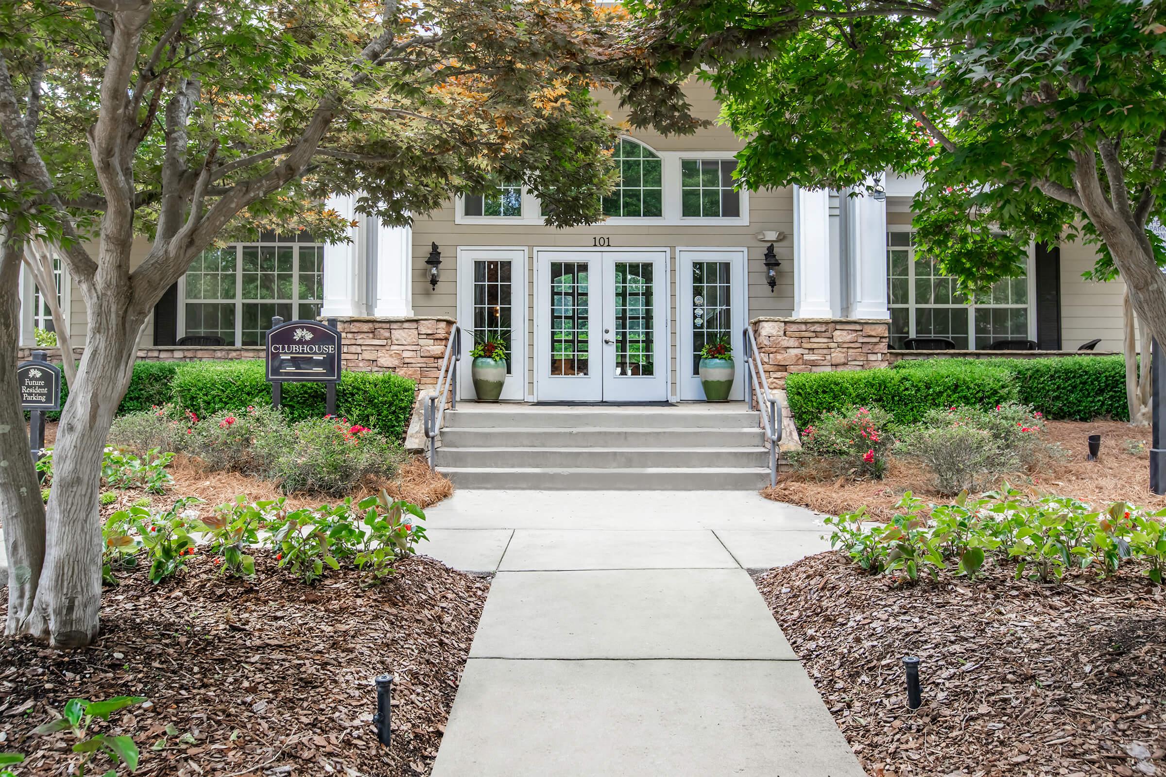Entrance of a clubhouse featuring stone and white pillars, surrounded by greenery and flower beds. A concrete pathway leads to double doors flanked by decorative potted plants. The surrounding area is well-maintained, with a welcoming atmosphere.
