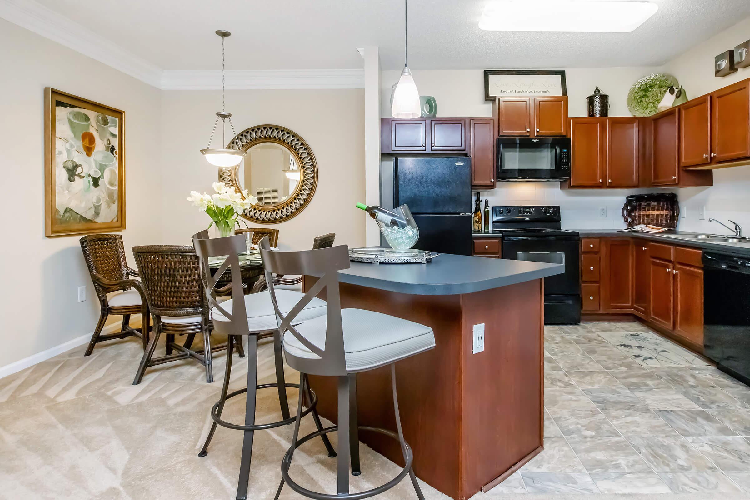 A modern kitchen featuring dark wood cabinets, a black refrigerator and stove, and a large island with bar stools. There is a round dining table set for four with decorative elements, and a piece of art on the wall. The floor has a patterned design, adding a stylish touch to the space.