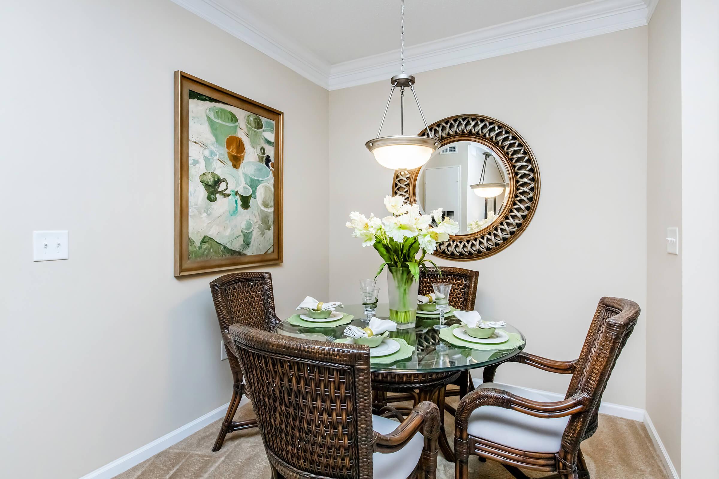 A dining area featuring a round glass table surrounded by four wicker chairs. The table is set with green plates and white napkins. A vase of white flowers is at the center. A large circular mirror and an abstract artwork hang on the wall, adding elegance to the neutral-colored space.