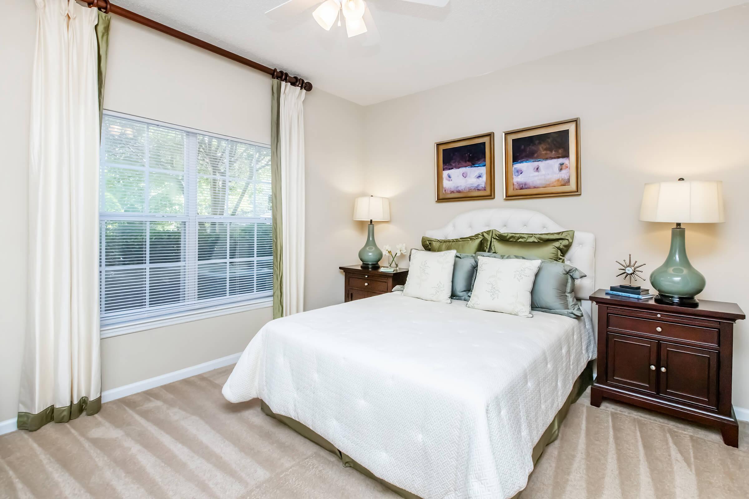 A cozy bedroom featuring a queen-sized bed with decorative pillows, two bedside tables with lamps, and two framed paintings above the bed. Natural light filters through a window adorned with sheer curtains, and a neutral color scheme creates a calm atmosphere.
