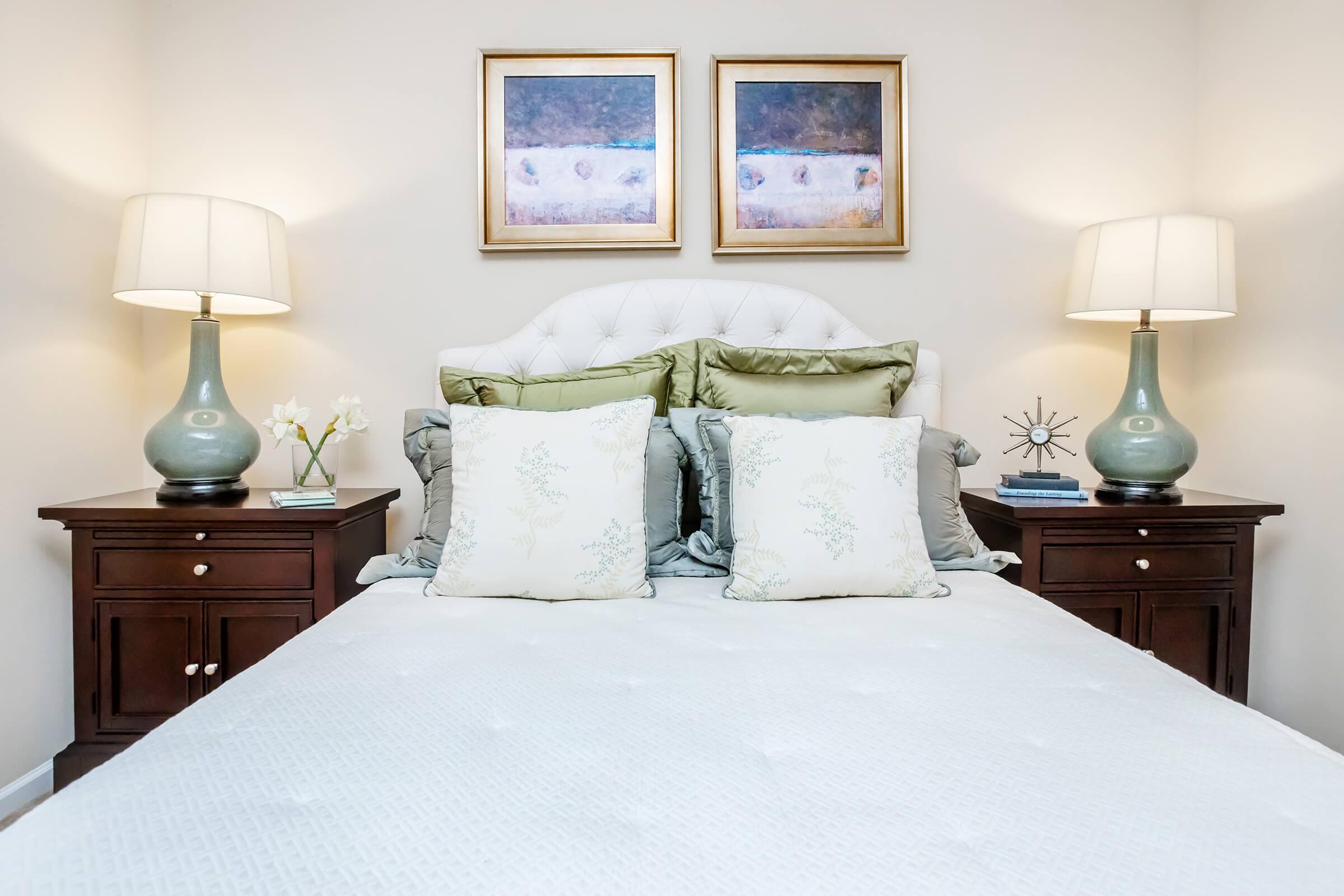 A neatly made bed with decorative pillows and a light-colored bedspread. On either side of the bed, there are wooden nightstands with lamps and small decorative items. Above the bed, two framed artworks hang on the wall, adding a touch of color to the serene bedroom decor.