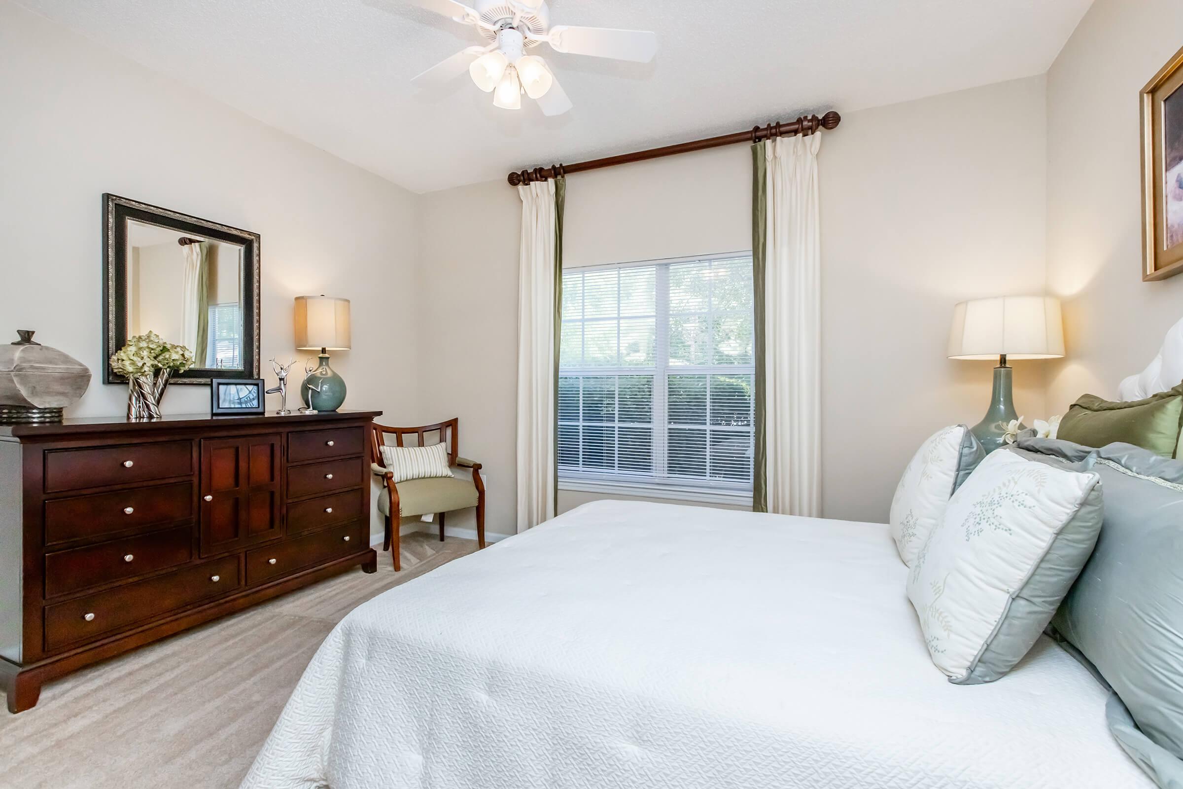 Cozy bedroom featuring a queen-sized bed with decorative pillows, a wooden dresser, a mirror, and bedside lamps. There's a chair by the window with sheer curtains allowing natural light in, creating a warm and inviting atmosphere. A ceiling fan is installed for added comfort.