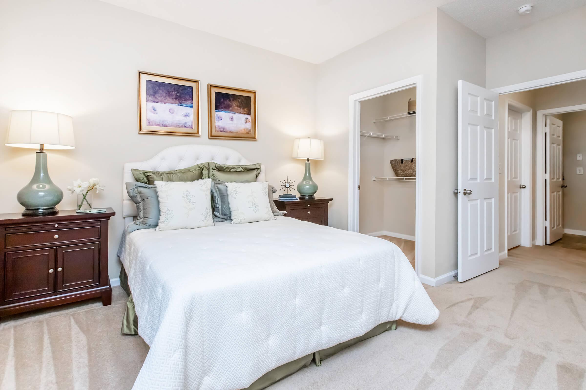 A cozy bedroom featuring a queen-sized bed with decorative pillows, bedside tables with lamps, and artwork on the walls. There is a closet with shelving and a doorway leading to a hallway. The room is softly lit, creating a warm and inviting atmosphere.