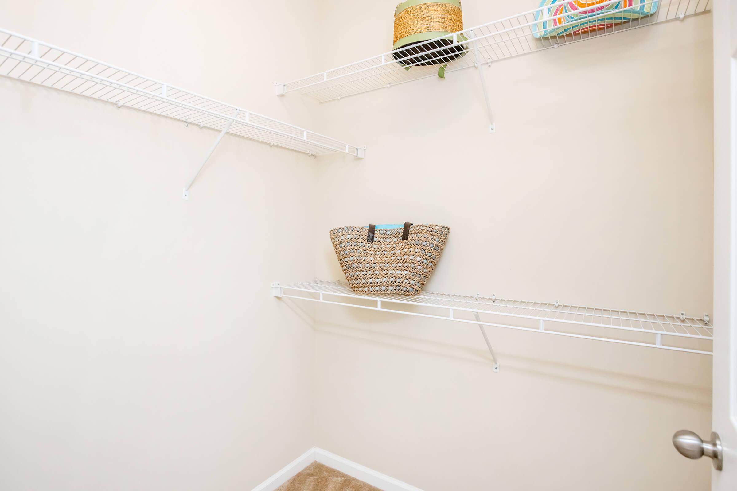 A clean and empty closet featuring white wire shelves. One shelf holds a woven basket, while a decorative container sits atop another shelf. The walls are painted a neutral color, and the floor is carpeted.